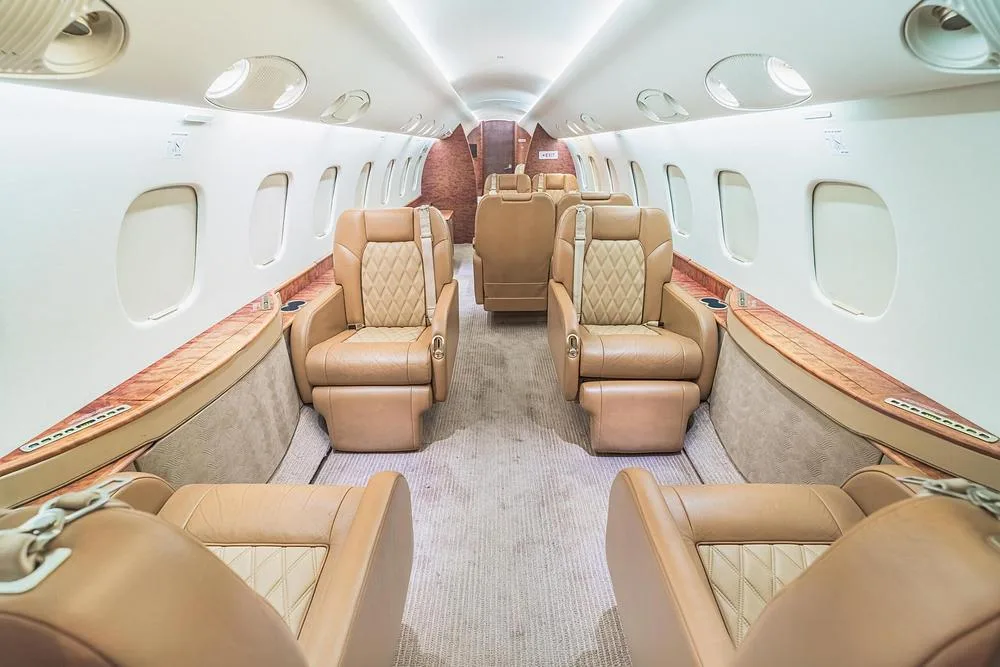 The luxurious cabin interior of a business jet features tan leather seats with diamond stitching and wood accents.