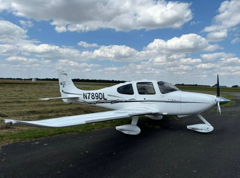 Side view of a white Cirrus SR22 aircraft with black accents and registration N789DL on a grassy airfield.