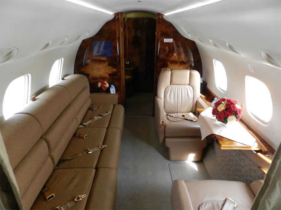 A private jet cabin interior showing a tan sofa on the left and a single tan leather seat with a table and flowers on the right.