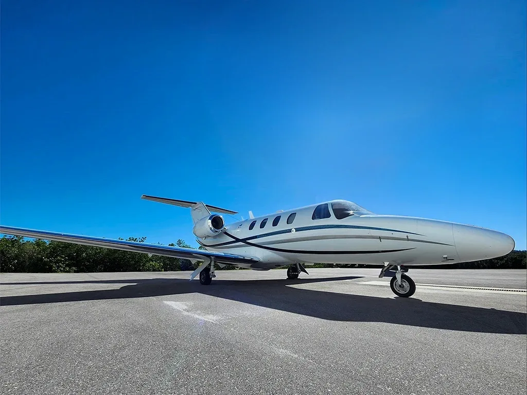 A white private jet with blue and grey stripes sits on an asphalt tarmac under a clear blue sky.