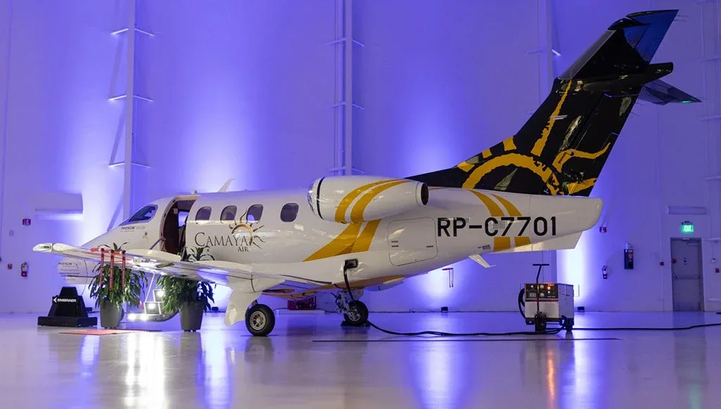 A white Embraer Phenom 300E with yellow and black Camaya Air livery parked inside a hangar with blue lighting.