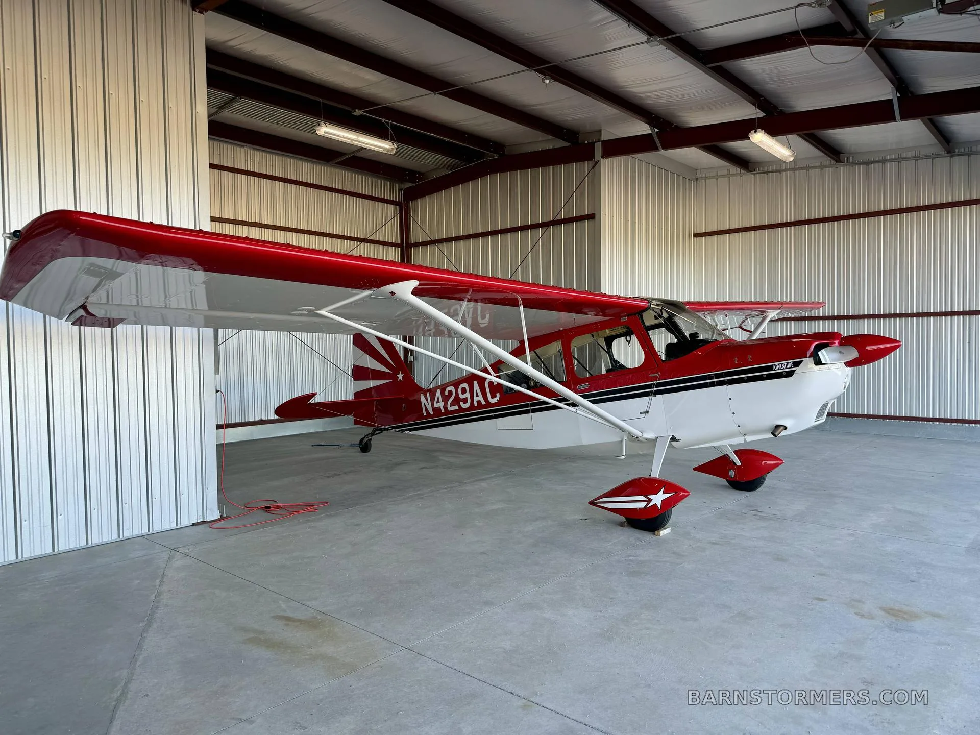 A red and white Adventure aircraft, N429AC, with a star on the wheel fairings, parked inside a hangar.