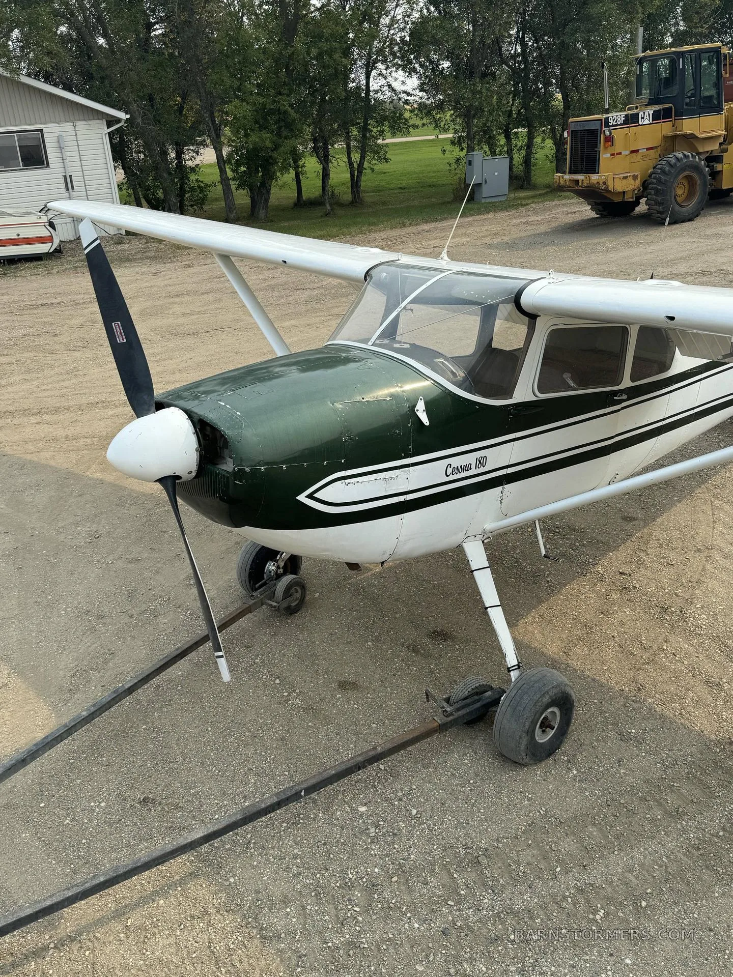 A front-left view of a white and green Cessna 180 aircraft parked on a gravel lot with heavy machinery in the background.