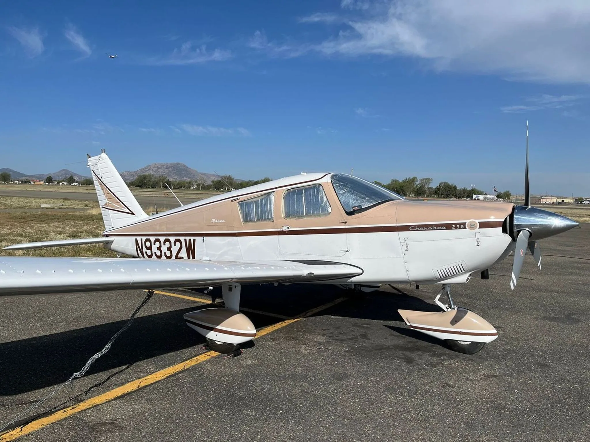 A Piper Cherokee 235, registration N9332W, in white and brown paint, parked on an airfield with mountains in the background.