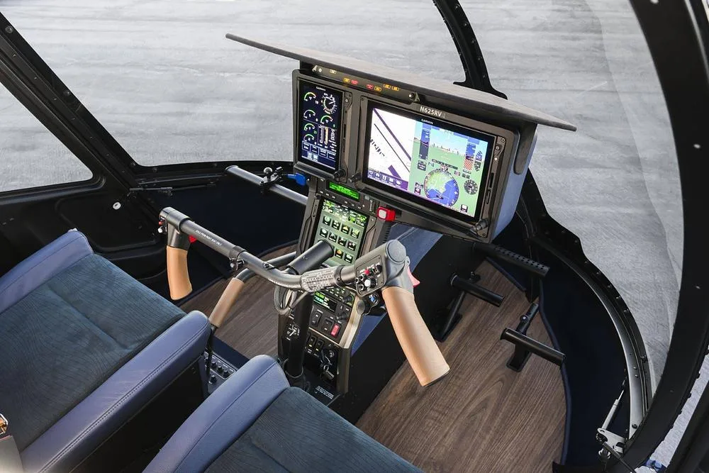 A wide view of a helicopter cockpit with dual digital screens, flight controls, and blue seats.
