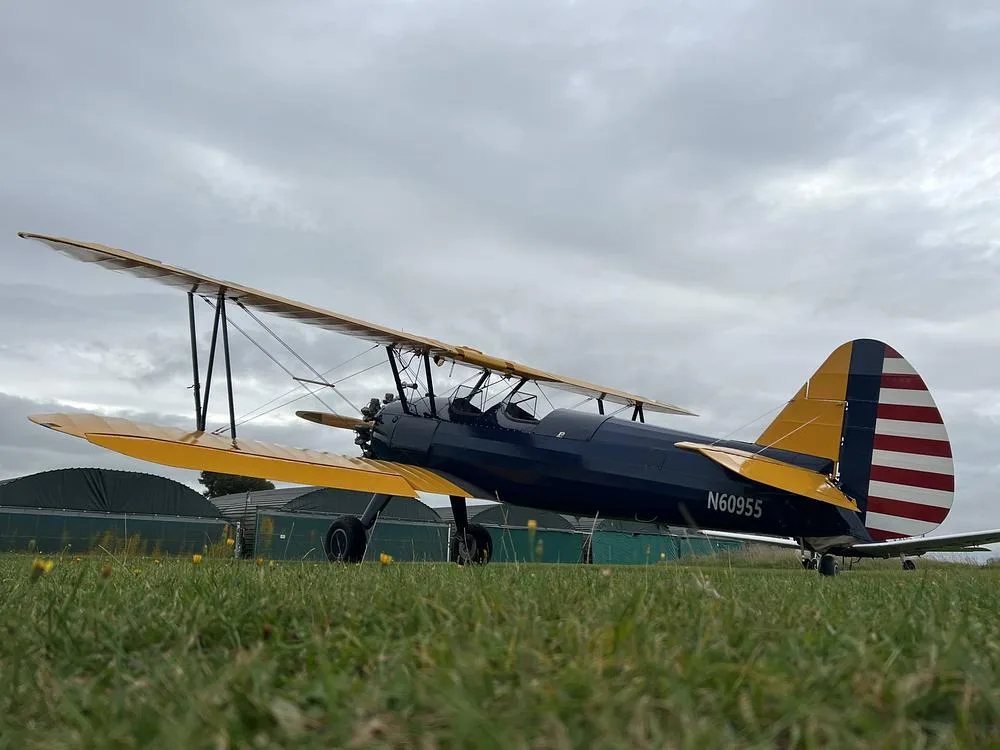 A low-angle side view of a blue and yellow biplane with N60955 registration parked on a grassy field with hangars in the background.