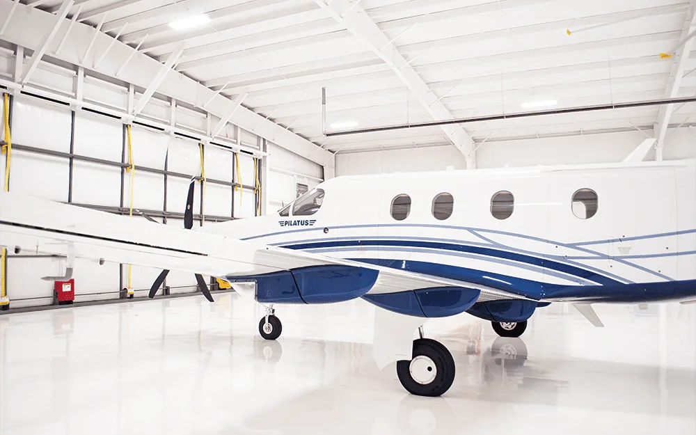 A side view of the white and blue Pilatus PC-12 aircraft, showing the landing gear and fuselage in a hangar.
