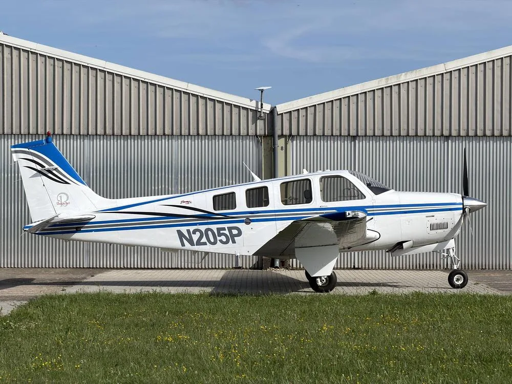 A white and blue Beechcraft Bonanza N205P parked on a paved surface next to a corrugated metal hangar.