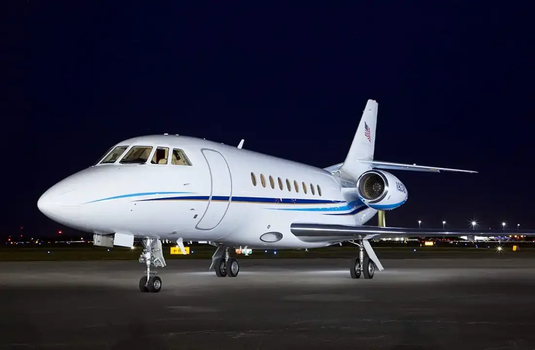 A white Dassault Falcon 900 business jet with blue stripes is parked on an airport tarmac at night.
