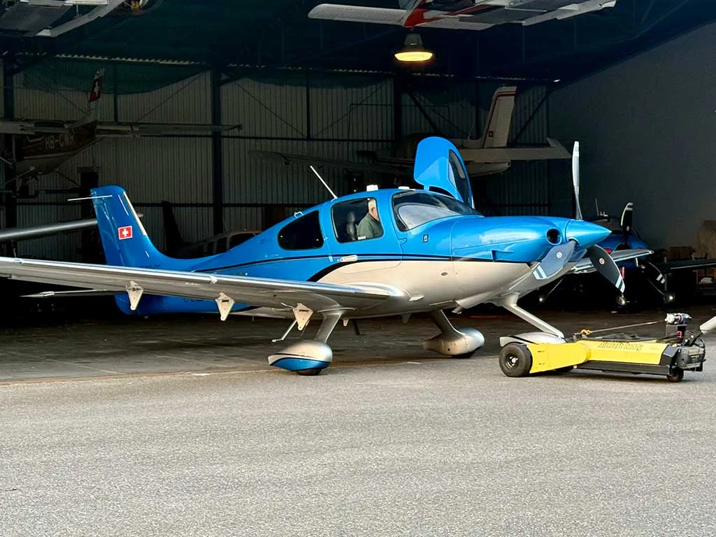 A blue and silver Cirrus SR22T aircraft with its door open, being towed out of a hangar.