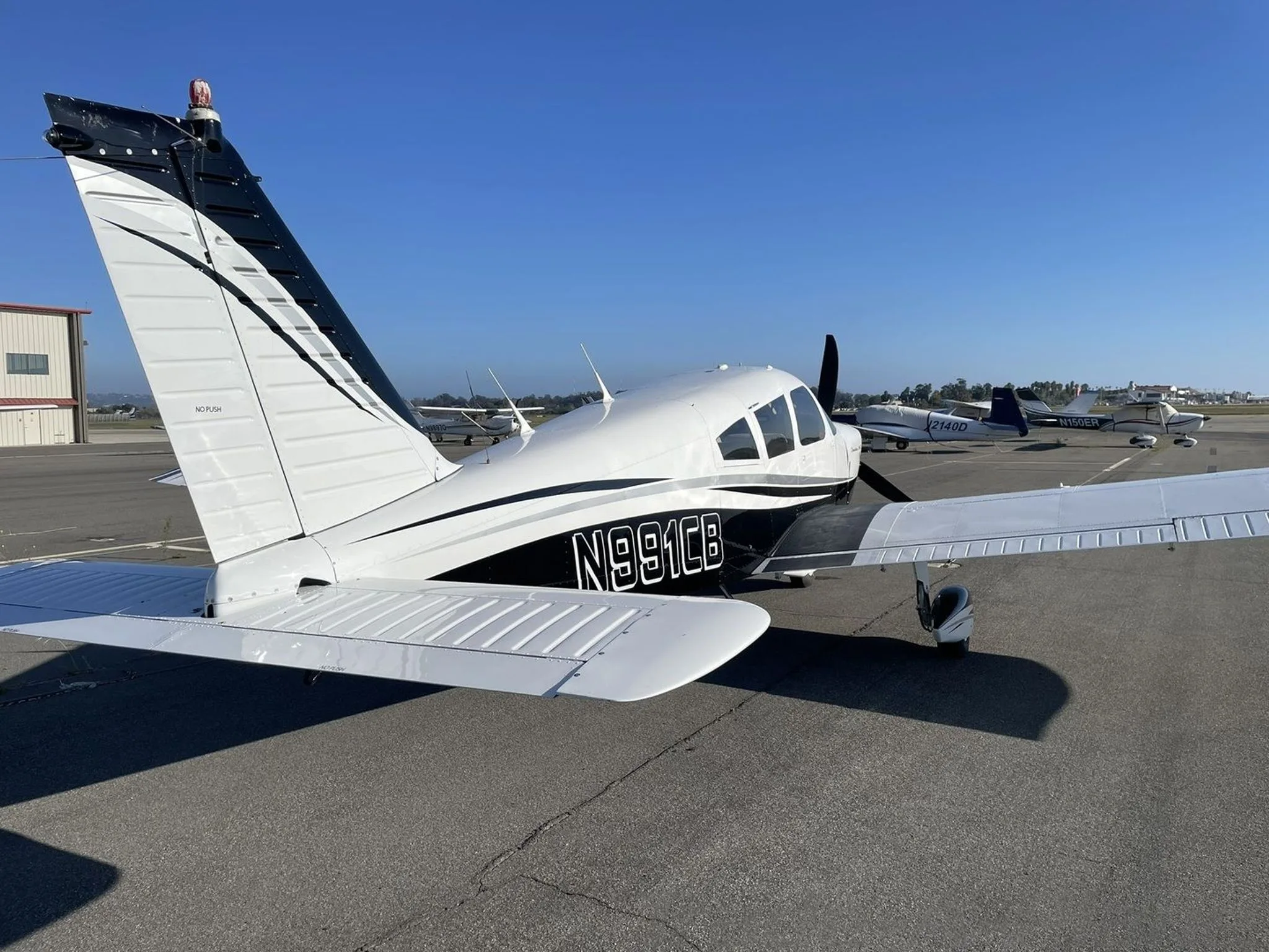 A rear-side view of the white and black Piper Cherokee 235 C aircraft N991CB parked on an airfield.