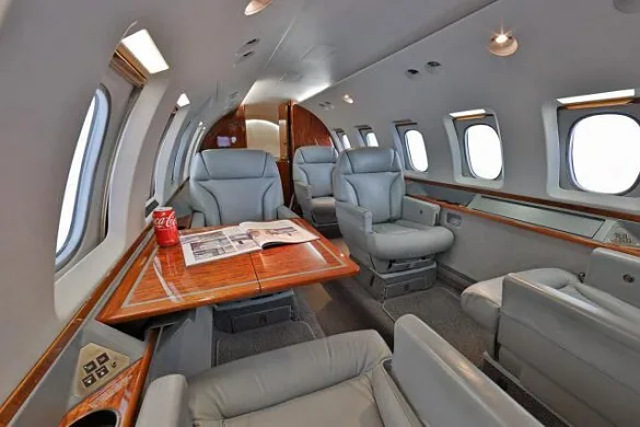 The luxurious interior cabin of a private jet with grey leather seats and a wooden table.