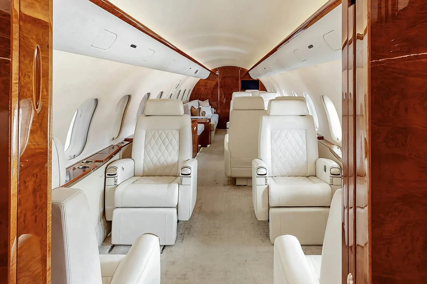 A view down the aisle of a business jet's cabin, showing multiple rows of light-colored quilted leather seats and polished wood paneling.