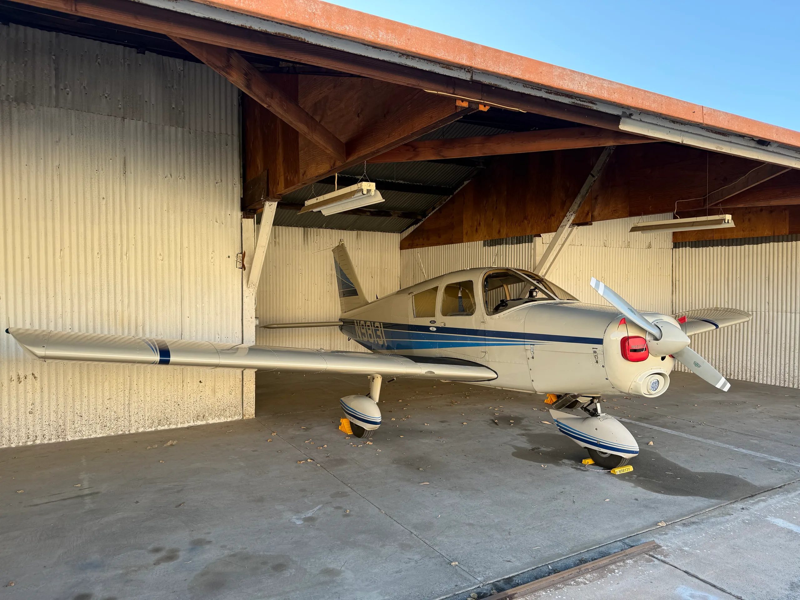 A white and blue Piper Cherokee aircraft (N98131) is parked under an open-sided hangar.