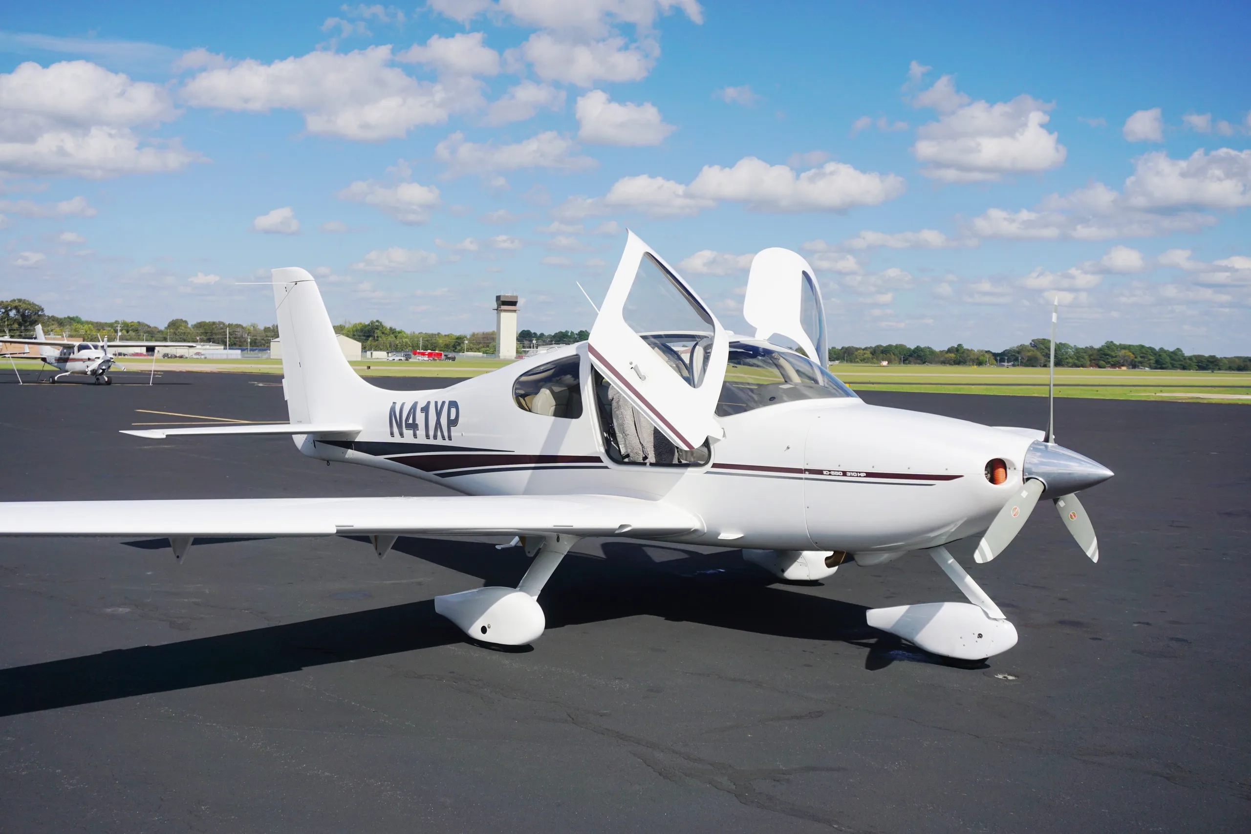 Side view of a white Cirrus SR22 with its gull-wing doors open, parked on an airport tarmac under a partly cloudy sky.