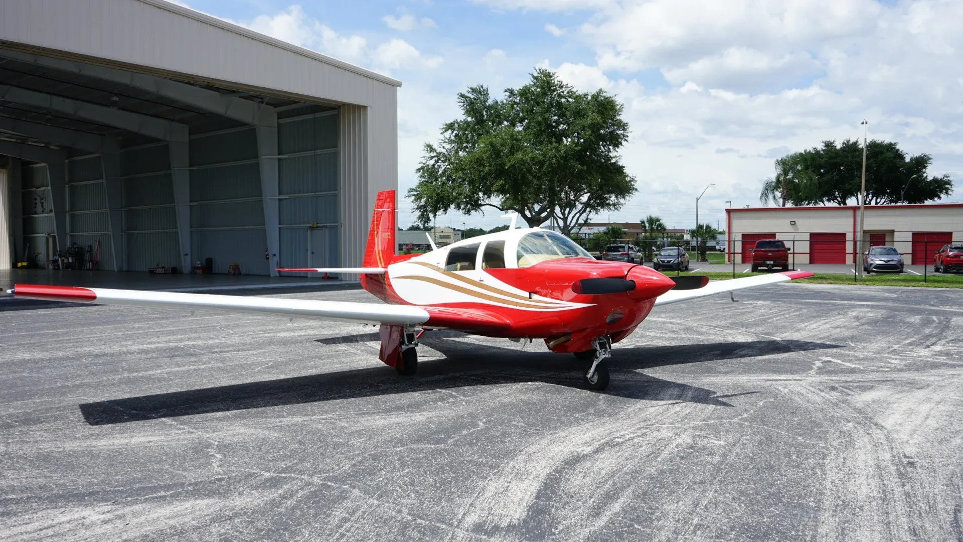 A red and white single-engine aircraft with gold stripes parked on an asphalt apron in front of an open hangar.