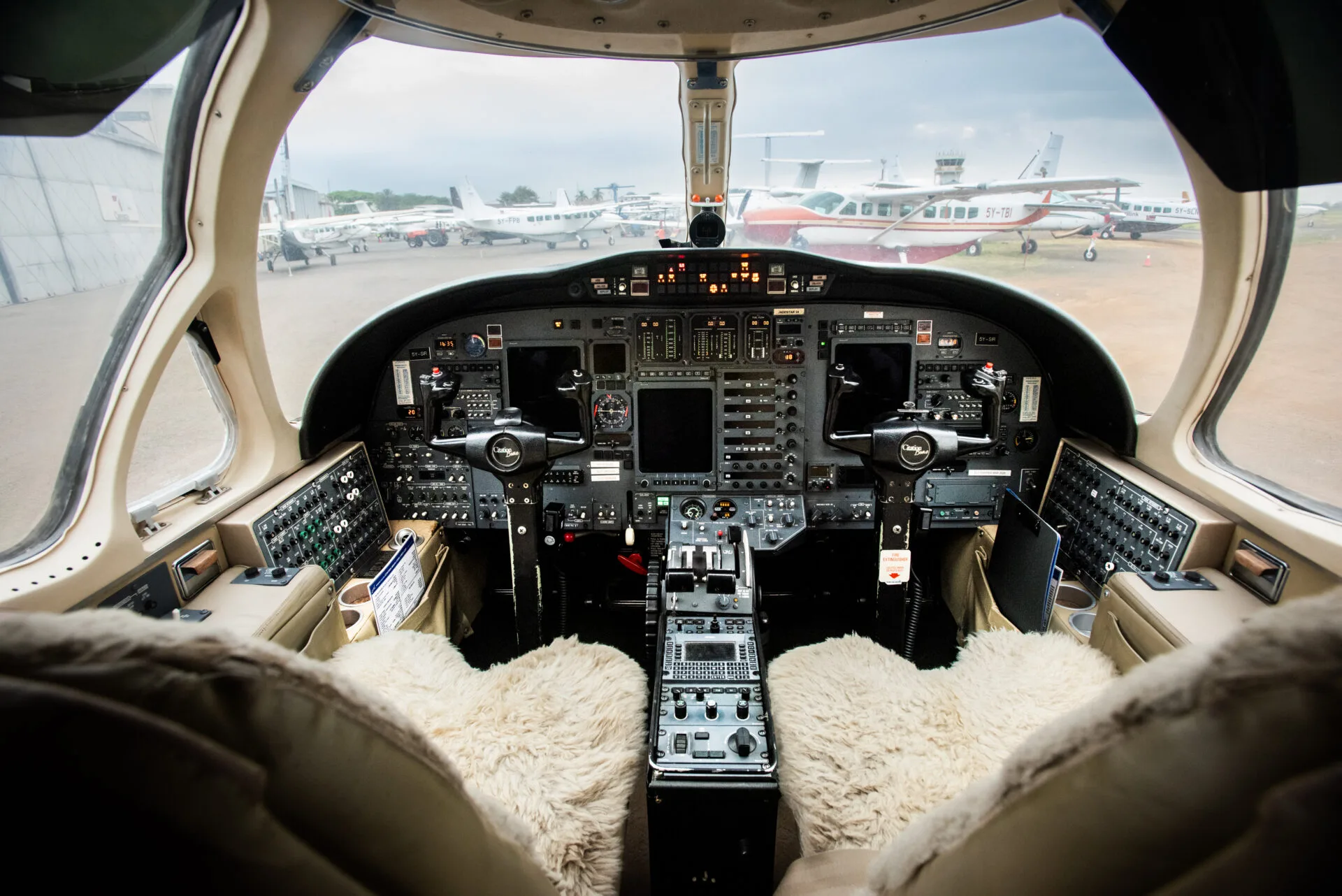 The cockpit of a business jet with fur seat covers, showing the instrument panel and other aircraft visible through the windshield.