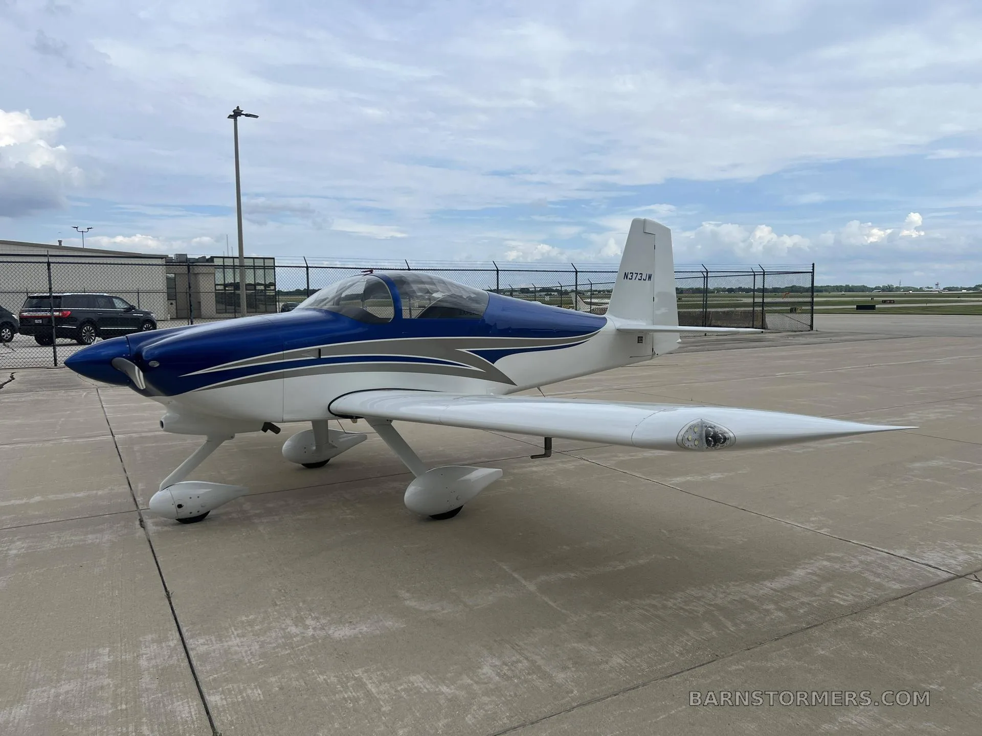 A white and blue single-engine aircraft, N373JW, with a bubble canopy, parked on a concrete tarmac under a cloudy sky.