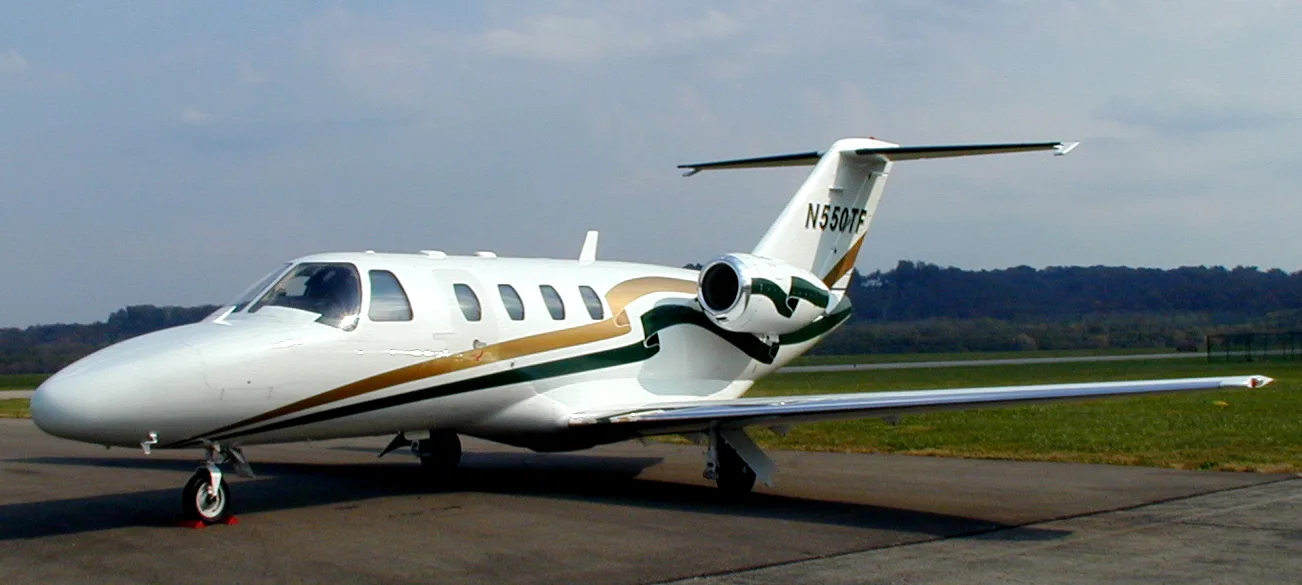 A white Cessna Citation business jet with green and gold stripes is parked on an airfield under a partly cloudy sky.