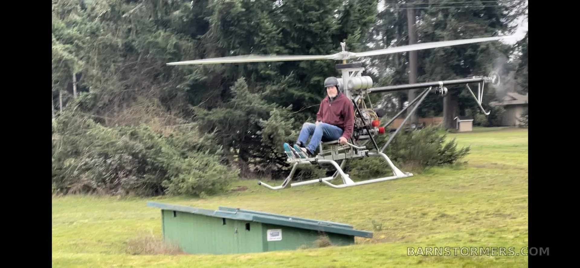 A pilot in a red jacket flies a single-seat ultralight helicopter low over a grassy field with trees in the background.