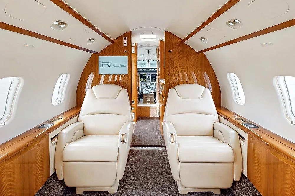 The aircraft cabin interior with cream seats and wood paneling, looking forward into the cockpit through an open door.