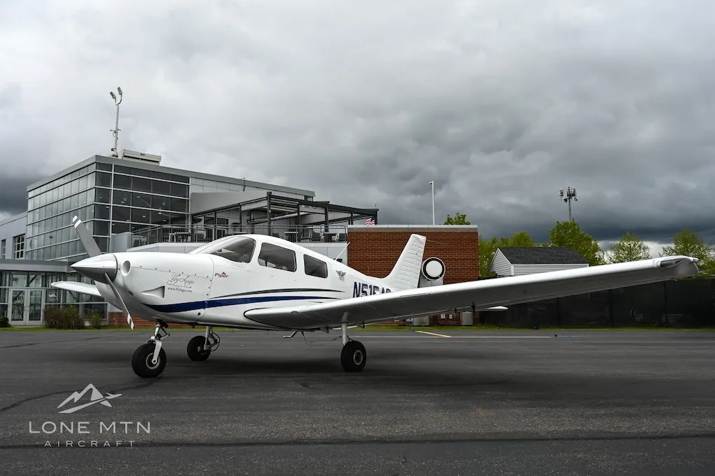 A white single-engine propeller aircraft with blue accents, registration N515AC, parked on an asphalt tarmac in front of a modern building.