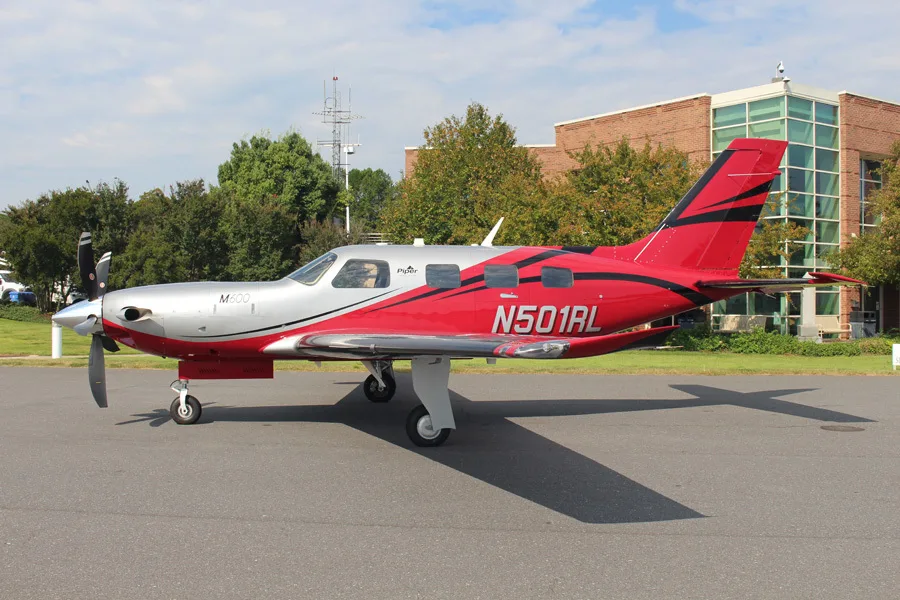 A Piper M600 aircraft, registration N501RL, in a red and silver paint scheme, parked on a tarmac with a building in the background.