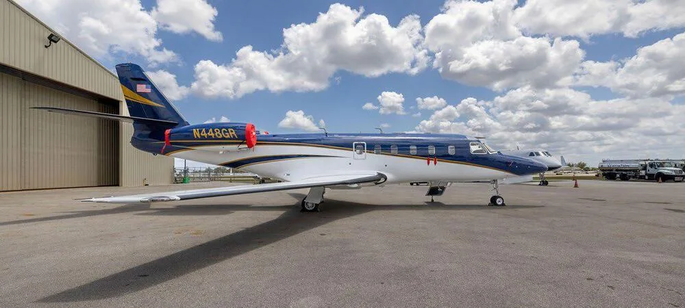 A blue and white business jet, N448GR, with gold stripes parked on an airfield next to a hangar under a cloudy sky.