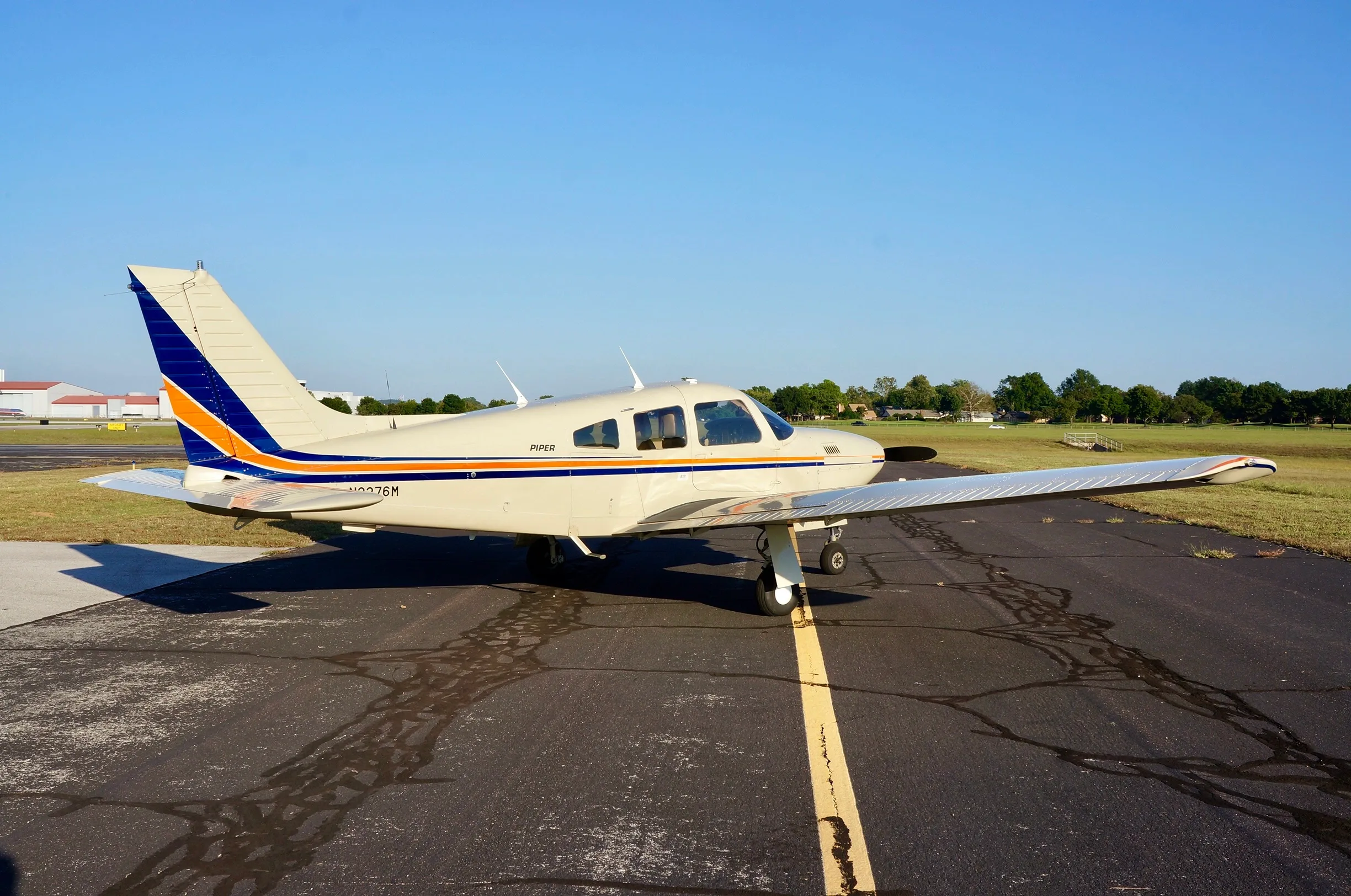 A cream-colored Piper aircraft, registration N2276M, with blue and orange stripes, viewed from the side on an airport tarmac.