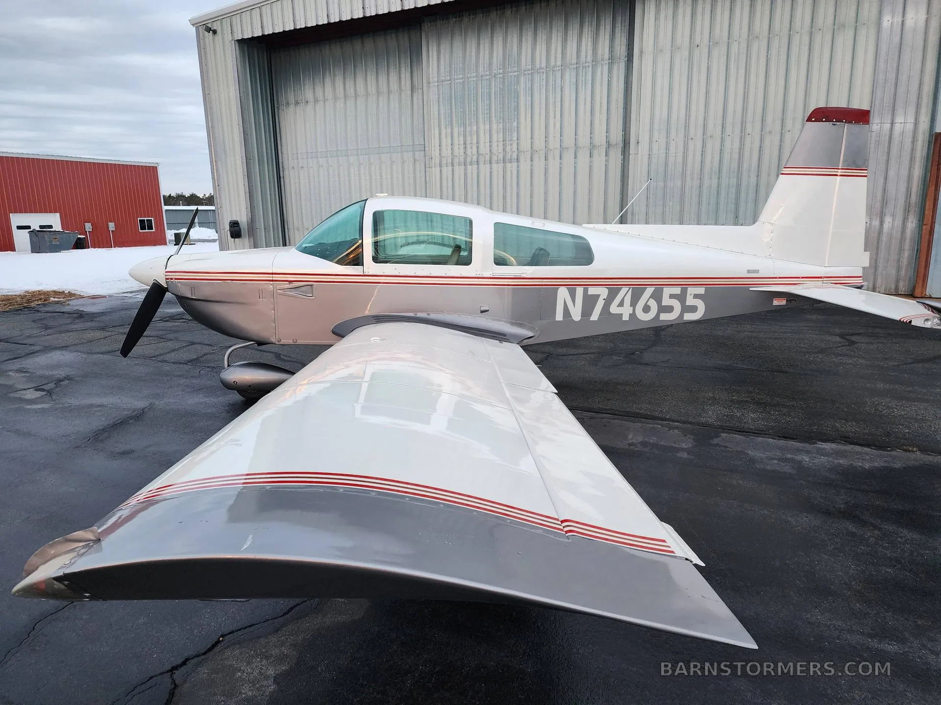 A white and silver Grumman AA-5 Traveler aircraft with red stripes, viewed from above and behind the left wing on a wet tarmac.