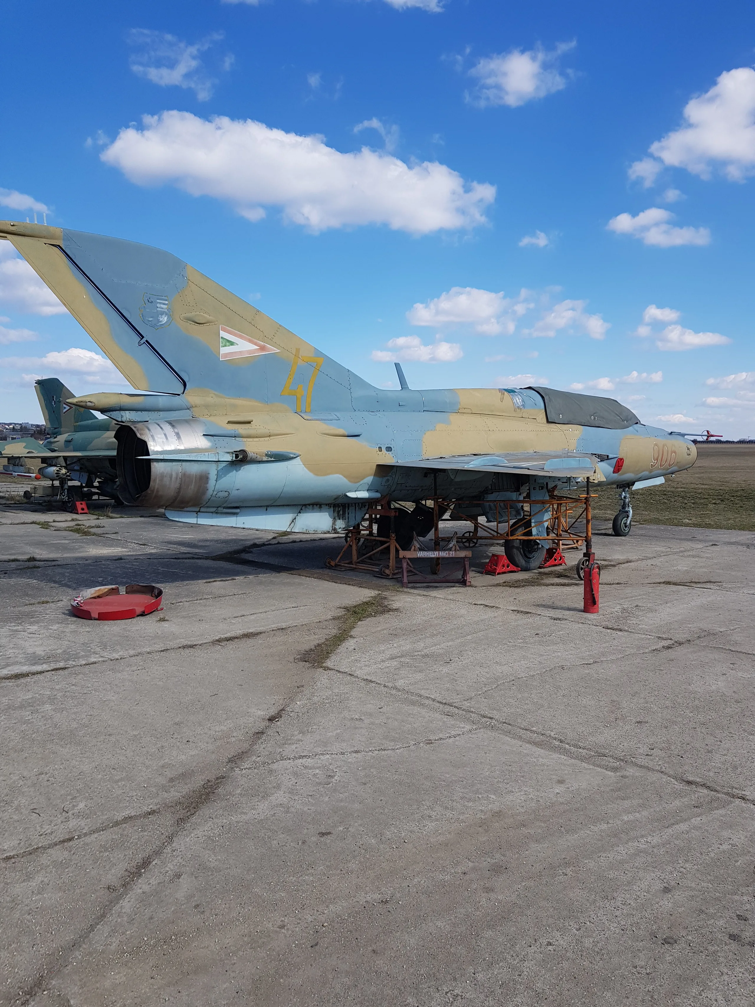 A MiG-21 fighter jet in blue and tan camouflage with Hungarian markings sits on a concrete airfield under a blue sky.