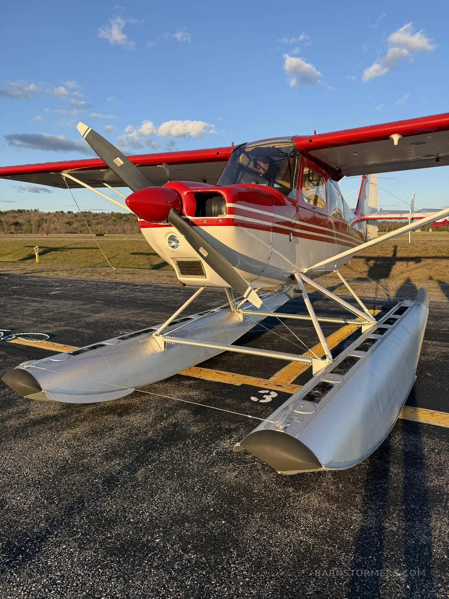 A red and white floatplane with a single propeller, parked on an asphalt runway under a blue sky.