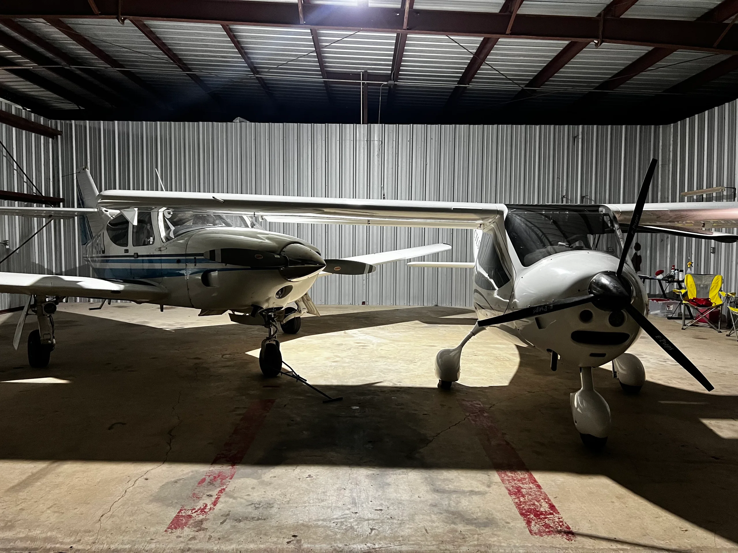 Two light aircraft, including a white Flight Design CTLS, parked side-by-side in a dimly lit hangar.