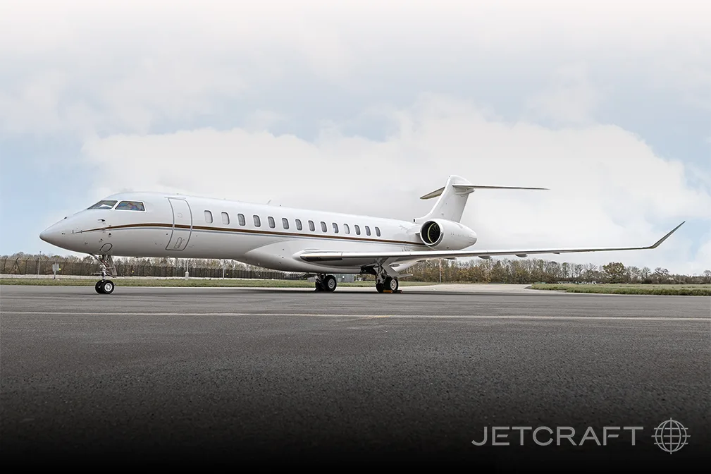 A white Bombardier Global business jet with a brown stripe on the fuselage is parked on an asphalt tarmac under a cloudy sky.