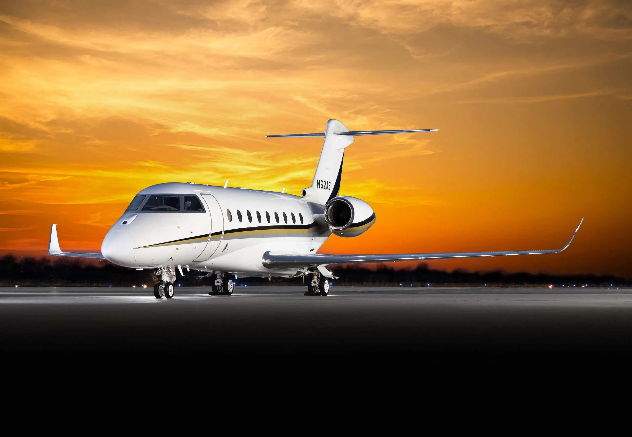 A white business jet with gold and black stripes on the tail, registration N62AE, parked on a tarmac at sunset.