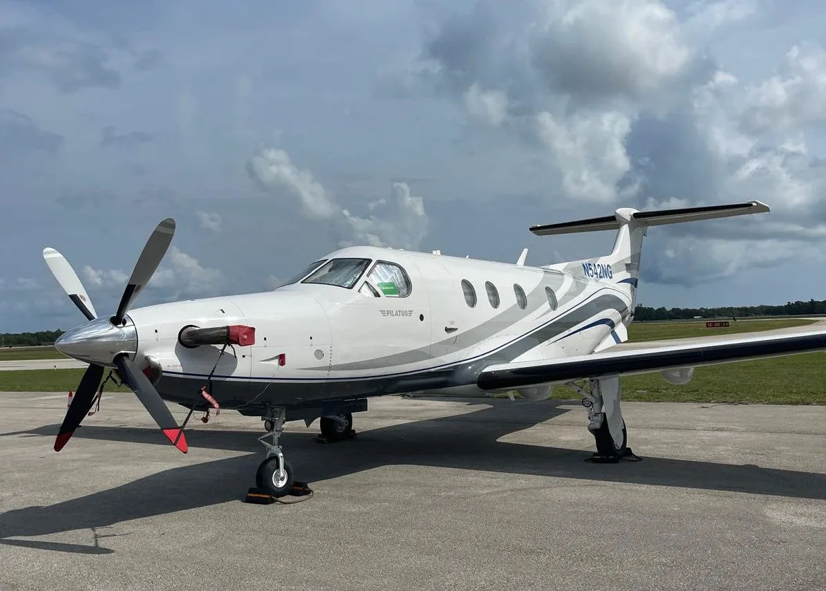 A white Pilatus PC-12 NG aircraft with blue and grey stripes, registration N542NG, parked on a tarmac under a cloudy sky.