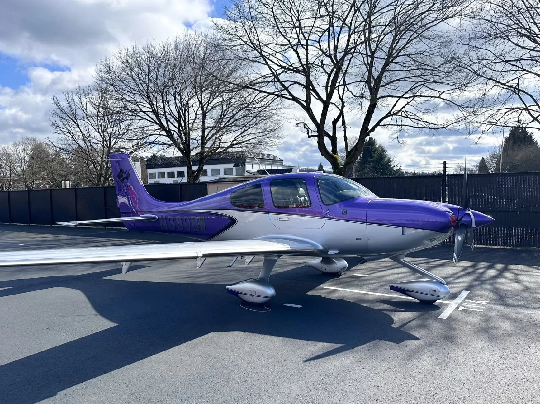 A side view of the purple and silver Cirrus SR22 aircraft, N380BW, parked on a paved surface with bare trees in the background.
