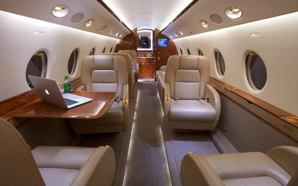 A luxurious aircraft cabin with tan leather seats, wood trim, and a laptop on a table.