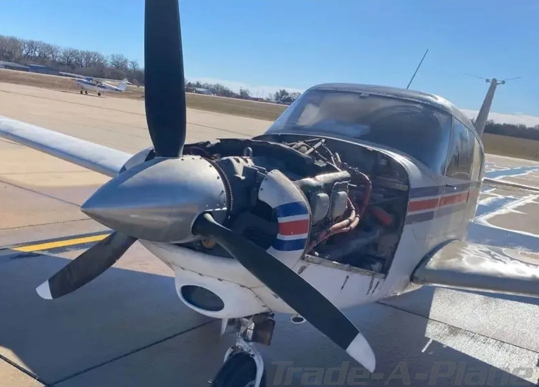 A white and grey single-engine propeller aircraft with its engine cowling open, parked on an asphalt runway on a sunny day.