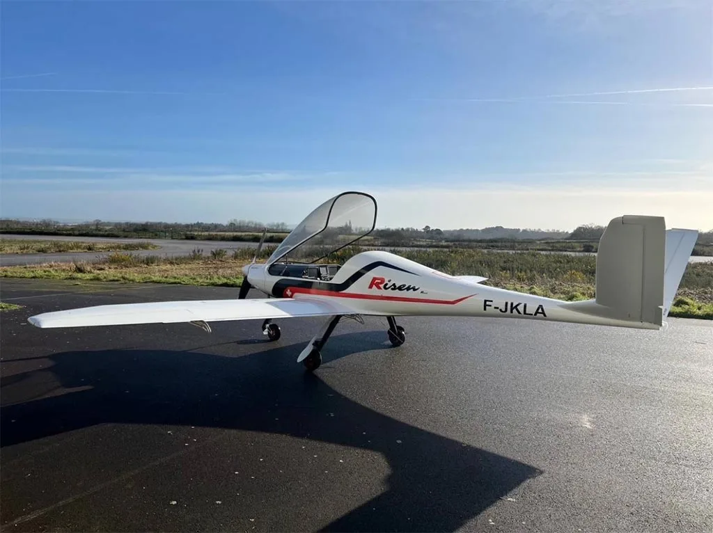 A white Risen aircraft with registration F-JKLA and an open canopy is parked on an airfield under a blue sky.