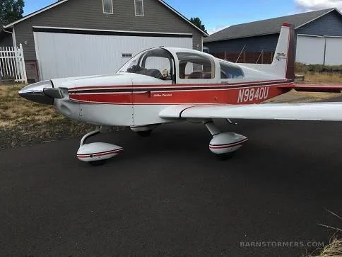 A white and red Grumman American AA-5 Traveler (N9840U) parked on asphalt in front of a hangar.