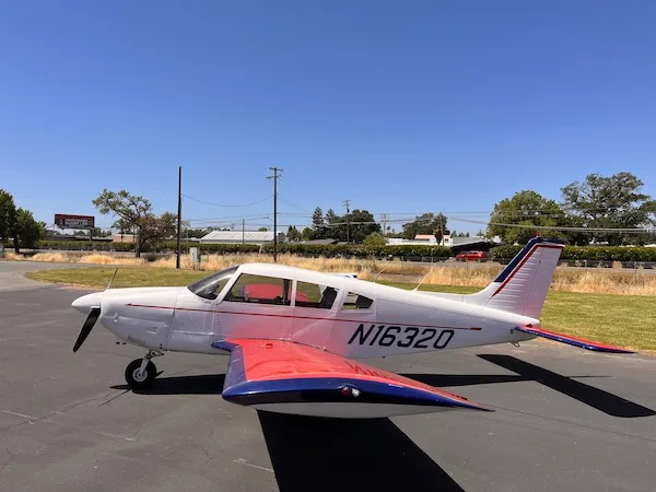 A white single-engine propeller aircraft with red wings and blue accents is parked on an asphalt surface under a clear blue sky.