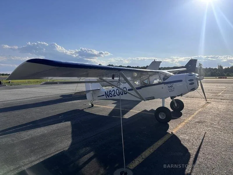 A white high-wing STOL aircraft with blue wing accents is parked on an asphalt ramp under a sunny sky.