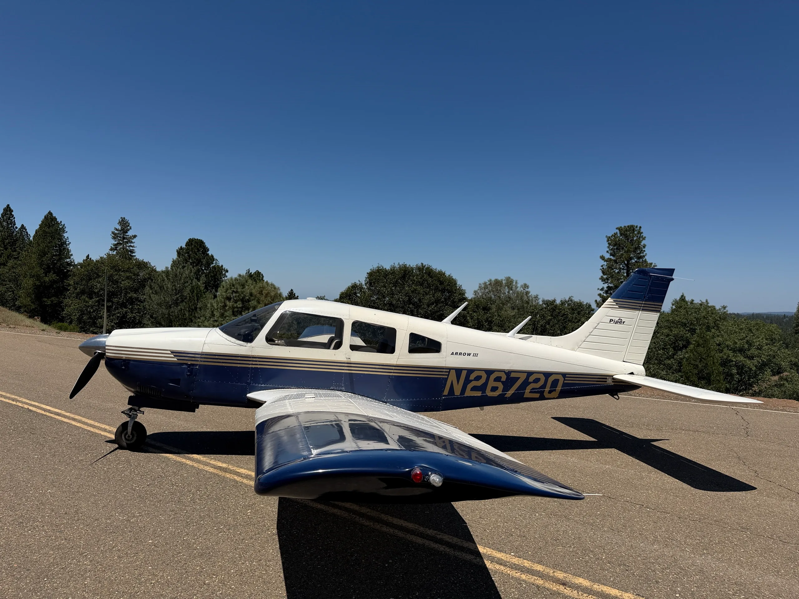 A white and blue Piper Arrow III aircraft, tail number N2672Q, parked on an asphalt surface with trees under a clear blue sky.