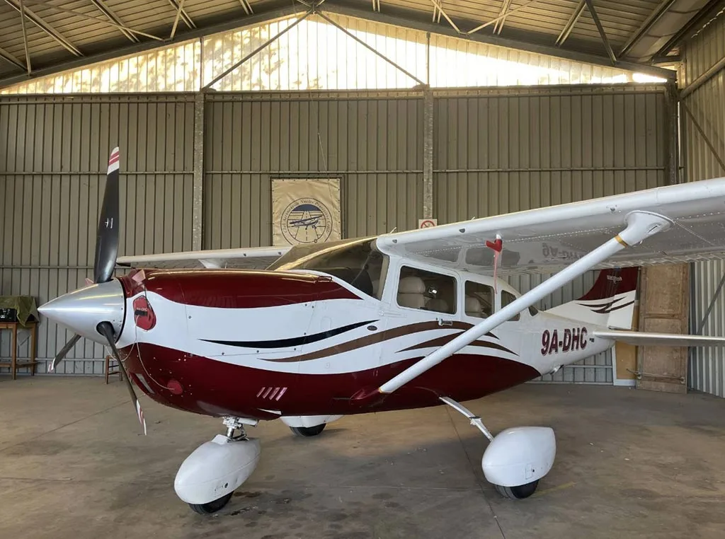 A red and white Cessna 206/210 aircraft with registration 9A-DHC parked in a hangar.