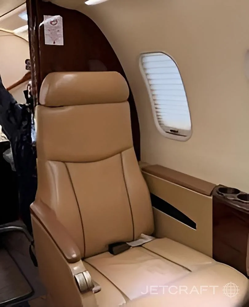 A single tan leather seat is positioned next to a window with a closed blind in an aircraft cabin.