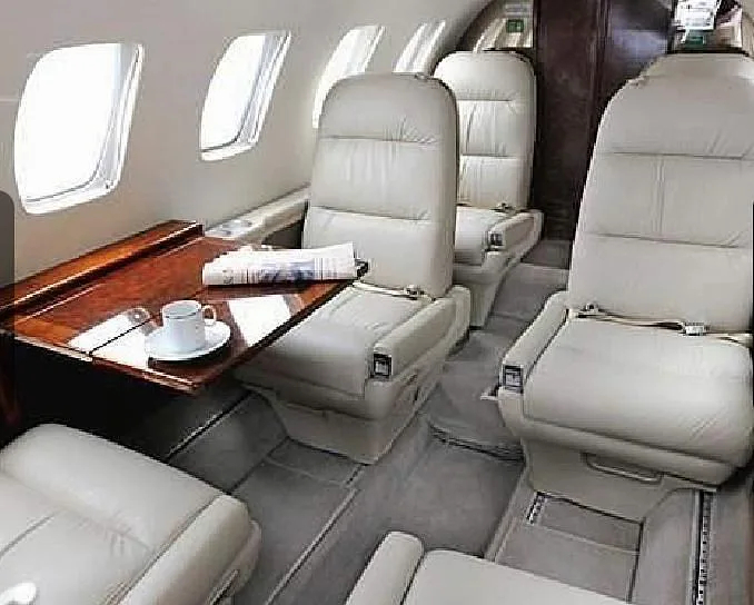 A close-up view of the aircraft cabin interior with light-colored leather seats, a wooden table with a newspaper and a cup, and windows.