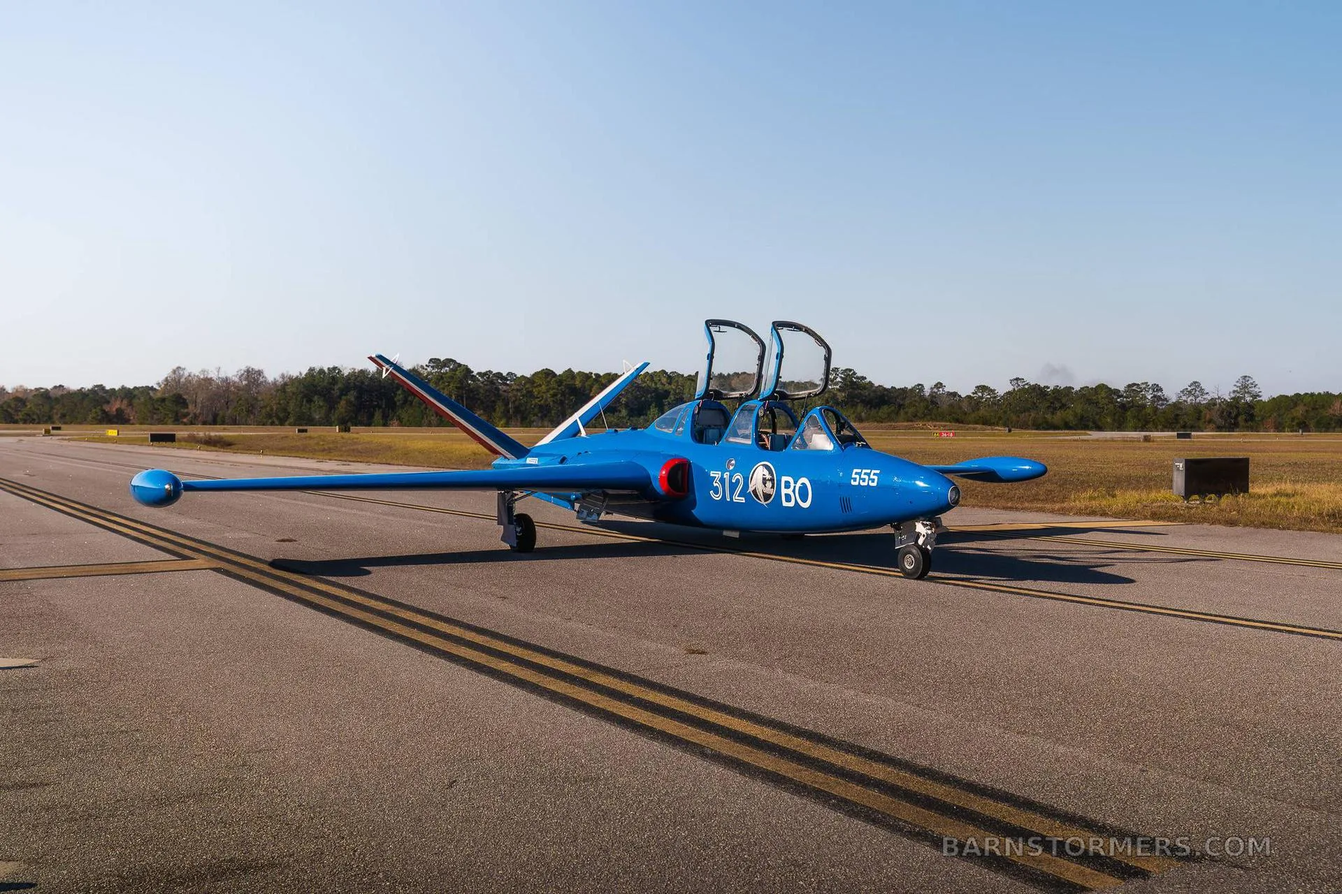 A blue Fouga CM.170 Magister jet aircraft with open canopies is parked on an asphalt runway.