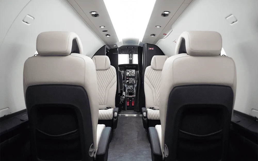A view down the aisle of the Pilatus PC-12 NGX cabin, showing the white and black quilted leather seats and the cockpit beyond.