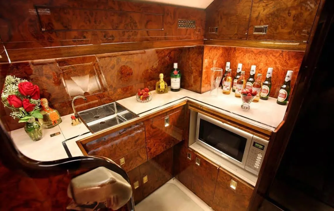 An aircraft galley with wood paneling, a double sink, microwave, and bottles of drinks and fruit on the counter.